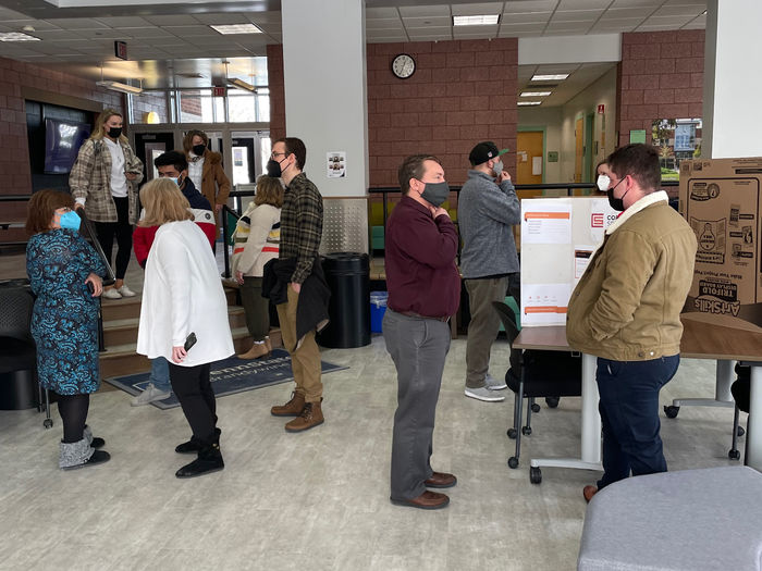 Students, faculty and staff gathered around posters in the lobby of a classroom building with large windows on Penn State Brandywine's campus.  