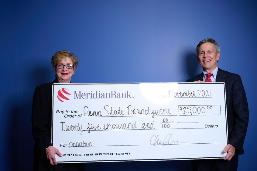 Meridian CEO Chris Annas with Penn State Brandywine Chancellor Marilyn Wells stand in front of a blue wall holding a large white check. 